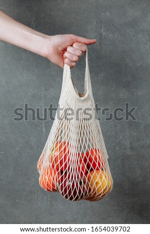 Mesh cotton shopping bag with mixed fruits in woman's hand. Plastic free, zero waste concept. Vertical shot. Gray textured background