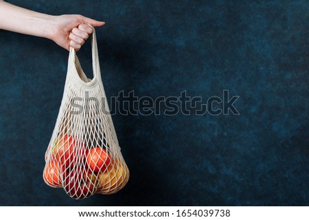Reusable mesh shopping bag with fruits in female hand. Modern reusable stores, zero waste concept. Dark blue textured background, copy space