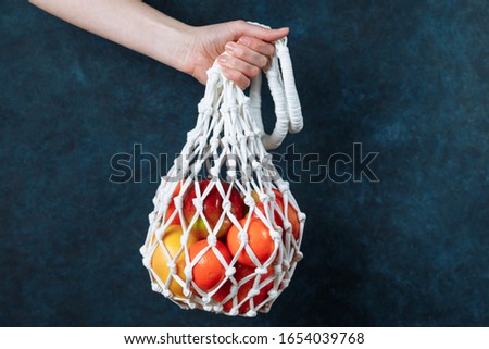 Female hand holds reusable mesh shopping bag of citrus fruits. Eco friendly and zero waste. Dark blue textured background