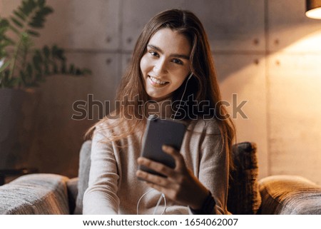 Smiling girl is sitting on sofa listening to music on headphones. Girls using smartphone for communication, entertainment. Millennial woman communicates in instant messengers, looks at camera, smile