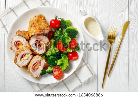 close-up of Chicken Cordon Bleu served on a white plate with broccoli, tomatoes, mustard Cream Sauce in a gravy boat on a table, horizontal view from above, flat lay, free space Royalty-Free Stock Photo #1654068886