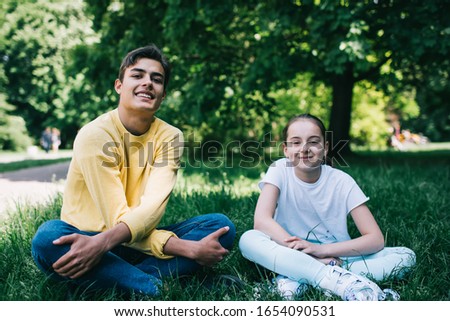 Happy teenager in casual outfit and girl in roller skates smiling and looking at camera while sitting with crossed legs on lawn on sunny day in park