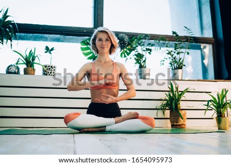 Smiling attractive blonde woman in yoga clothing sitting in lotus position on yoga mat surrounded by potted plants and looking at camera