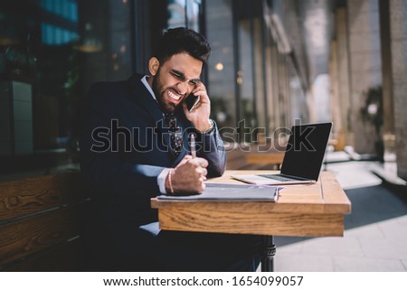 Annoyed furious businessman dressed in black suit white shirt tie surfing netbook making call on cellphone punching on wooden table in city cafe in summer 
