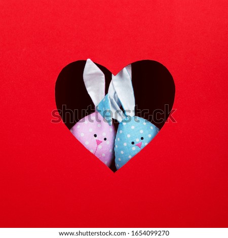 The concept of spring, happy Easter, eared pink and blue rabbit handmade, peek out of a hole in the shape of a heart on red background. Copy space.