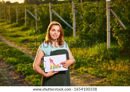 Woman farmer working in fruit garden and shows the level of crop growth using infographics. Biologist inspector examines raspberries bushes