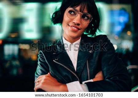 Pretty hipster girl in stylish eyeglasses dreaming about funny nightlife and smiling at metropolitan urban setting, carefree young woman dressed in stylish outfit standing on blurred background