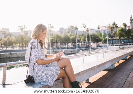 Side view of lost female traveler with digital camera sitting on long bench with legs crossed and looking for direction on mobile navigator on background with promenade and river