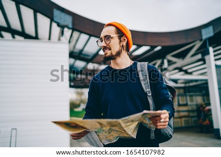 Happy Caucasian male tourist in stylish glasses smiling during travel sightseeing for exploring city streets, good looking backpacker holding paper map for search location direction and walking Royalty-Free Stock Photo #1654107982