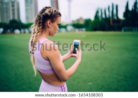 Side view of young sportive female in fitness wear standing on stadium and taking picture of football field on mobile phone