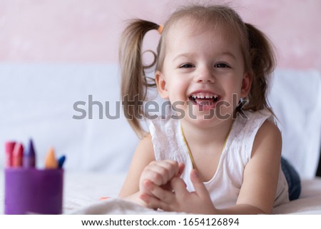 Beautiful girl draws with pencils in bed. Little girl two years paints coloring book lying on the bed. Little baby with a ponytail smiling while lying on the bed. Pretty girl paints with pencils and