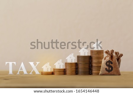 Mini house on rows of rising coins, US dollar bags, word tax on wood table. House or housing, land value, finance tax concept: depicts ad valorem tax on the value of the property all.