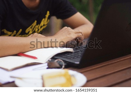 An Asian man wears a black T-shirt working and using a notebook computer in a beautiful garden in a coffee shop.