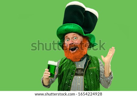 Pint of beer. Inviting. Excited leprechaun in green suit with red beard on green background. Funny portrait of man ready to party. Saint Patrick day, human emotions, celebration, traditional holidays.