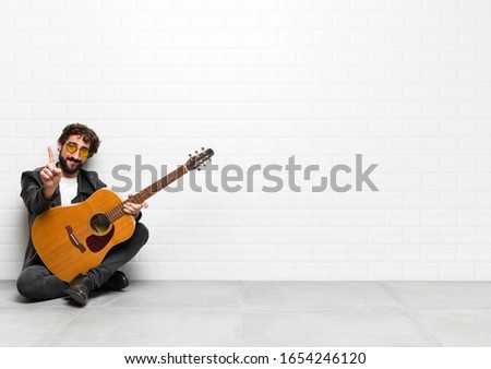 young musician man smiling and looking friendly, showing number one or first with hand forward, counting down with a guitar, rock and roll concept