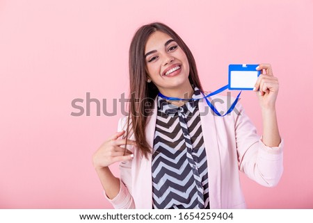 Beautiful success woman shows on camera her badge and smiling over pink wall. Young attractive woman holding blank artist lanyard or badge in hand with metal piece. Plastic pass concept.