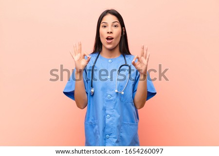 young latin nurse feeling shocked, amazed and surprised, showing approval making okay sign with both hands against pink wall