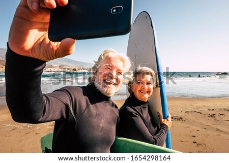 couple of pensioners seniors taking a selfie together at the beach with their wetsuits and surfboards - mature people learning surfing