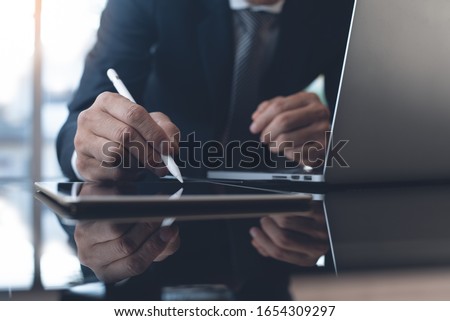 Businessman in black suit working in modern office. Business man with stylus pen signing on digital tablet screen, reviewing business report with laptop computer on glass table, close up