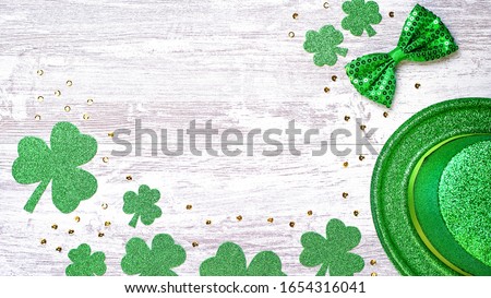 Happy Saint Patrick's Day greeting card with traditional symbols, shamrock, green attire. Green hat, bow tie, St Patricks Day shamrocks, golden confetti on white wooden background, copy space