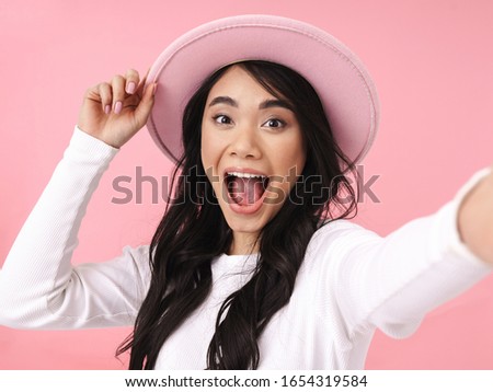 Cheerful pretty young asian woman taking a selfie while standing isolated over pink background, wearing hat