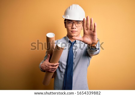 Young handsome chinese architect man wearing glasses and safety helmet holding blueprints with open hand doing stop sign with serious and confident expression, defense gesture