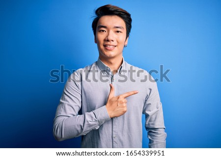 Young handsome chinese man wearing casual shirt standing over isolated blue background cheerful with a smile on face pointing with hand and finger up to the side with happy and natural expression