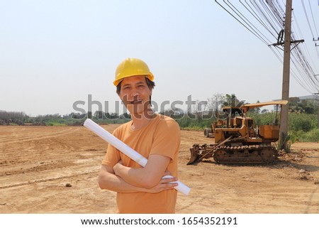 Male civil engineer or architect wear the yellow helmet and cross one arm with project drafts while in hand standing on background of front loader engineering construction vehicle at the work area.
