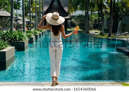 Female brunette in hat (no face), with resort style clothes white color on her slim body, enjoying holiday in tropic garden. She standing near blue swimming pool, holding glass of fresh orange drink   Royalty-Free Stock Photo #1654360066