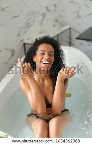 Young afro american curly haired girl taking bath with flowers and wearing black bra and panties. Concept of home bathroom underwear photo session and hygiene.