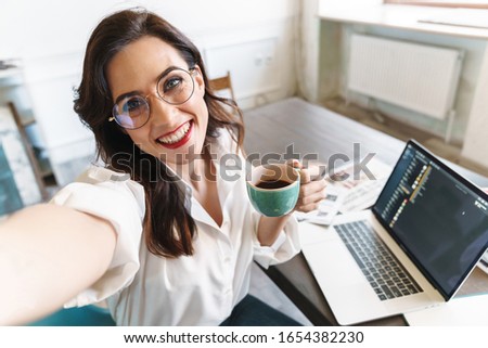 Attractive cheerful young brunette businesswoman sitting at the cafe table with laptop computer indoors, taking a selfie, holding cup of coffee