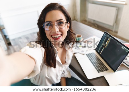 Attractive cheerful young brunette businesswoman sitting at the cafe table with laptop computer indoors, taking a selfie