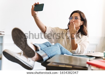 Lovely cheerful young brunette woman relaxing at the table in the cafe indoors, taking a selfie