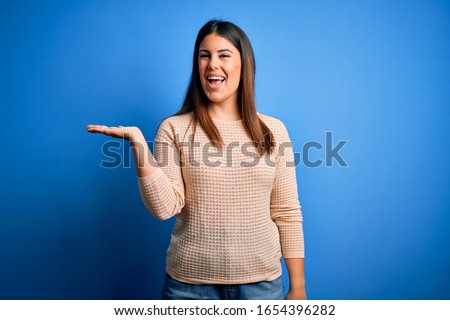 Young beautiful woman wearing casual sweater over blue background smiling cheerful presenting and pointing with palm of hand looking at the camera.