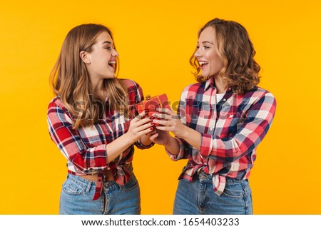 Image of two young beautiful girls wearing plaid shirts smiling and holding gift box isolated over yellow background