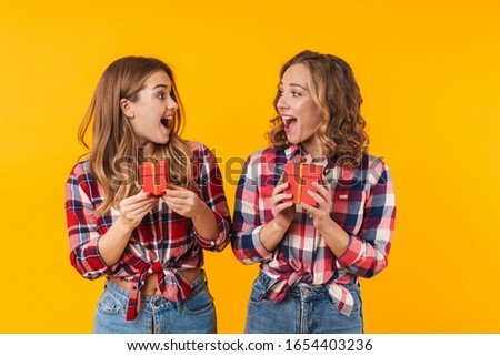 Image of two young beautiful girls wearing plaid shirts smiling and holding gift boxes isolated over yellow background