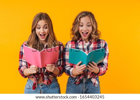 Image of two young beautiful girls wearing plaid shirts smiling and holding diary books isolated over yellow background