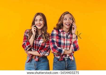 Image of two young beautiful girls wearing plaid shirts smiling and using cellphones isolated over yellow background