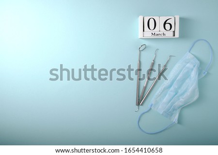 Metal dental tools, wooden calendar with with the date March 6, mask on the face, on a blue background