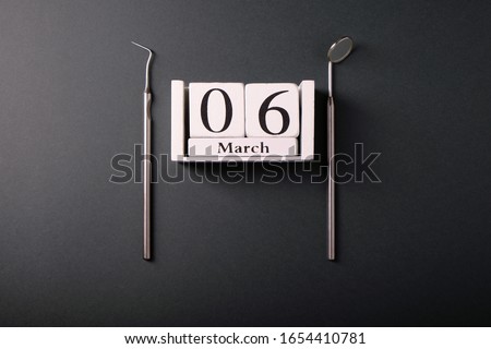 Metal dental tools, wooden calendar with with the date March 6, on a dark background, top view