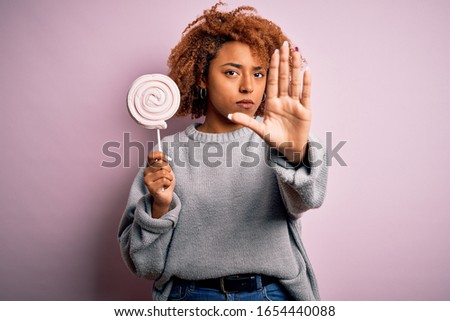 Young beautiful African American afro woman with curly hair eating sweet candy with open hand doing stop sign with serious and confident expression, defense gesture