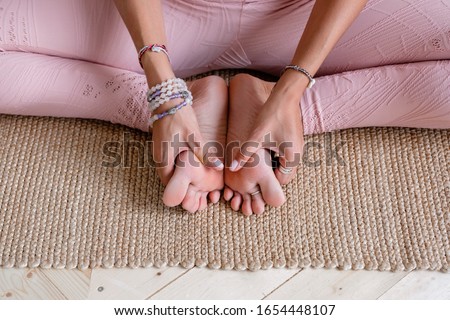 An unidentified young slim woman in a pink suit is doing a foot massage and sitting in the lotus position. Young woman yoga teacher does an advanced lotus position during yoga class