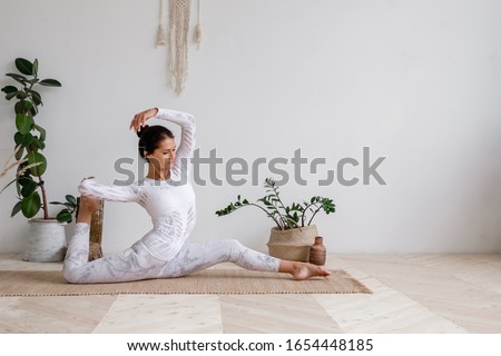 Slim pretty young brunette woman in a tight light suit makes hanumanasana sitting on a rug on the floor surrounded by houseplants on a white background. Advanced amateur concept. Advertising space