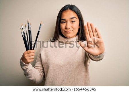 Young beautiful artist asian woman holding paint brushes standing over white background with open hand doing stop sign with serious and confident expression, defense gesture