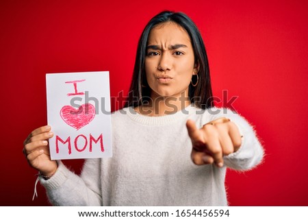 Young beautiful woman holding paper with love mom message celebrating mothers day pointing with finger to the camera and to you, hand sign, positive and confident gesture from the front