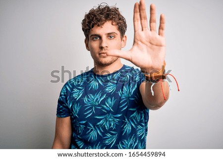Young blond handsome man on vacation with curly hair wearing casual summer t-shirt doing stop sing with palm of the hand. Warning expression with negative and serious gesture on the face.