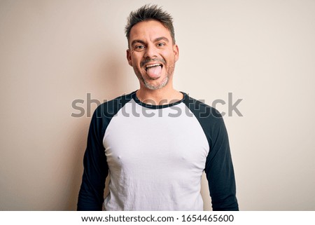 Young handsome man wearing casual t-shirt standing over isolated white background sticking tongue out happy with funny expression. Emotion concept.
