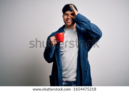 Young blond man with beard and blue eyes wearing pajama drinking cup of coffee with happy face smiling doing ok sign with hand on eye looking through fingers