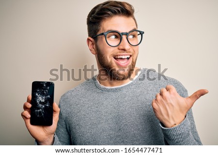 Young blond man with beard and blue eyes wearing glasses holding broken smartphone pointing and showing with thumb up to the side with happy face smiling