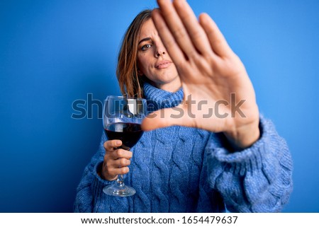 Young beautiful brunette woman drinking glass of red wine over isolated blue background with open hand doing stop sign with serious and confident expression, defense gesture